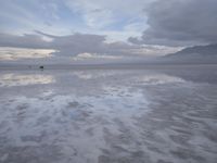 a lonely beach with cloudy skies in the distance, reflecting water on the sand and a building on the shore of the other side