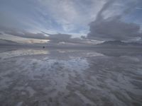 a lonely beach with cloudy skies in the distance, reflecting water on the sand and a building on the shore of the other side