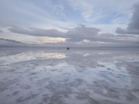 a lonely beach with cloudy skies in the distance, reflecting water on the sand and a building on the shore of the other side