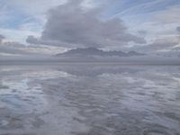 a lonely beach with cloudy skies in the distance, reflecting water on the sand and a building on the shore of the other side