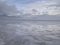 a lonely beach with cloudy skies in the distance, reflecting water on the sand and a building on the shore of the other side