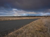 a wet, lonely road leading to a grassy plain under a cloudy sky with a dark gray sky