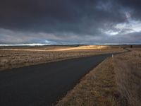 a wet, lonely road leading to a grassy plain under a cloudy sky with a dark gray sky