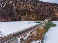 a long bridge spanning over a large frozen lake with some snow on the ground and hills in the background