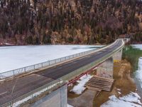 a long bridge spanning over a large frozen lake with some snow on the ground and hills in the background