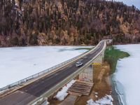 a long bridge spanning over a large frozen lake with some snow on the ground and hills in the background