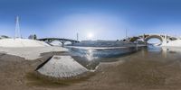 a fisheye view of a river and a bridge on the shore with a sand hill in the background