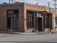 the motorcycle is stopped near the brick building on the street corner beside the curb, and with one leg visible