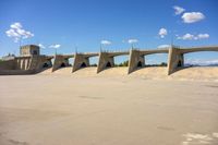 cement bridge overpass on dirt field under cloudy blue sky, with water flowing below