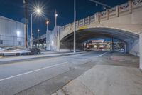 the street under the bridge is empty and the lights are on and cars drive under it