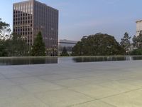 the walkway at the park leads to a lake and buildings in the background as evening falls