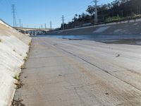 water running along side a cement wall under a bridge and road light with electrical wires