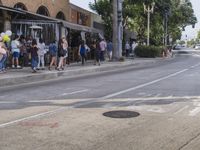 people wait on the sidewalk at an intersection with shops on either side of the street