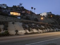 an empty highway with light at night on the side of a mountain ridge and buildings behind it