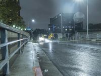 a wet street at night with cars passing by in the rain, and a person standing on a sidewalk, under a streetlight