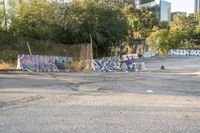 a large parking lot filled with lots of graffiti and trash cans near trees and a wire fence