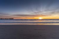 the sun setting behind a pier and ocean waves on a beach with footprints in the sand