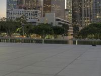 the walkway at the park leads to a lake and buildings in the background as evening falls