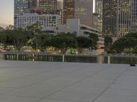 the walkway at the park leads to a lake and buildings in the background as evening falls