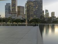 the walkway at the park leads to a lake and buildings in the background as evening falls