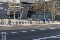 a long row of bicycles on the sidewalk by a building and trees and a fence