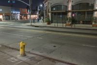 a yellow fire hydrant in an empty street by a building at night with stop lights