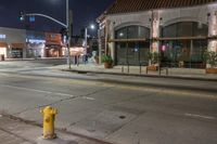 a yellow fire hydrant in an empty street by a building at night with stop lights