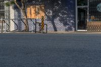 an empty street with three wooden chairs in front of a storefront window with a few people nearby