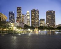 the large city skyline is lit up at dusk from a park across from the water