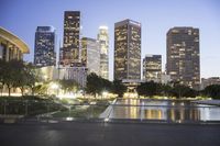 the large city skyline is lit up at dusk from a park across from the water