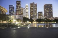 the large city skyline is lit up at dusk from a park across from the water