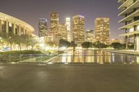 the large city skyline is lit up at dusk from a park across from the water