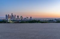 a city skyline sits in the distance at sunset by the road side, with a park bench