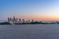 a city skyline sits in the distance at sunset by the road side, with a park bench