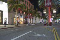 palm trees in an intersection next to a busy city street at night time with people walking on the sidewalk