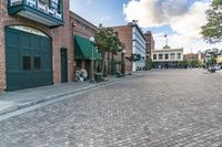 a brick city street with people sitting on the curb and buildings lining the street with green shutters