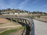 a curved metal bridge with buildings near by in the background in a hilly area with dry grass and dirt