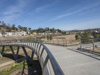 a curved metal bridge with buildings near by in the background in a hilly area with dry grass and dirt