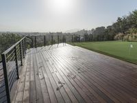 a wooden deck with grass and a fenced in view of a golf course with hills and trees