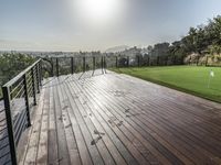 a wooden deck with grass and a fenced in view of a golf course with hills and trees