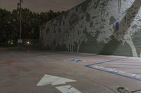 an empty parking lot with a wall painted with trees and benches in the background, a tree with many leaves, blue lines painted on it