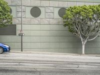 a blue car is driving down the road near a building on a windy day with small trees in front