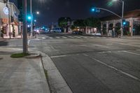 an empty street lit up at night, with cars parked on the curb and green traffic lights hanging above it