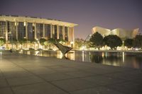 there is a bench and a lake in front of a large building at night time