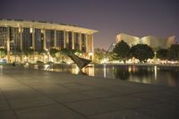 there is a bench and a lake in front of a large building at night time