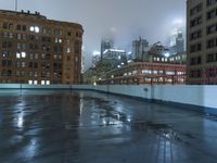 a wet roof at night next to tall buildings and skyscrapers with reflections in the puddle