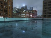 a wet roof at night next to tall buildings and skyscrapers with reflections in the puddle