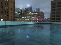 a wet roof at night next to tall buildings and skyscrapers with reflections in the puddle