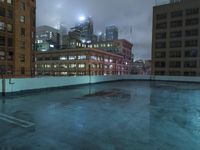 a wet roof at night next to tall buildings and skyscrapers with reflections in the puddle
