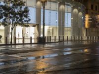 the sidewalk has some rain on it near some buildings at night with one pedestrian walking away